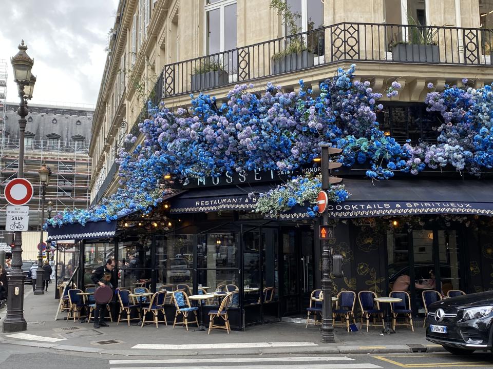 A French café on a street corner with outside seating and blue flowers on the awning