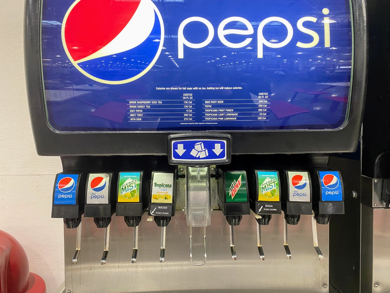 Six Pepsi products at the soda fountain at the Costco food court, one machine with eight selections and ice, white wall and trash can in the background
