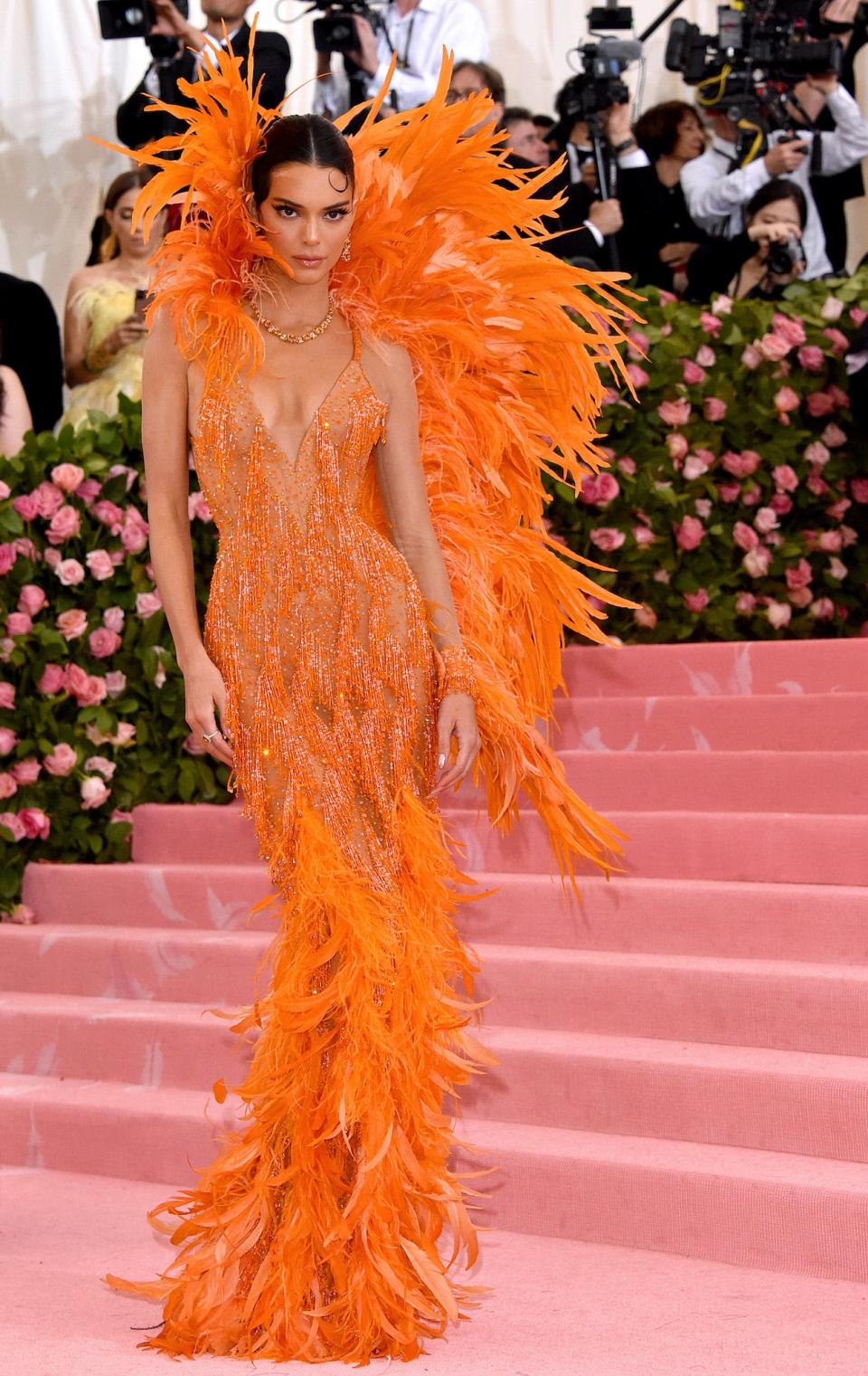 Kendall Jenner poses on the Met Gala stairs in an orange feathered dress with a giant feathered collar.