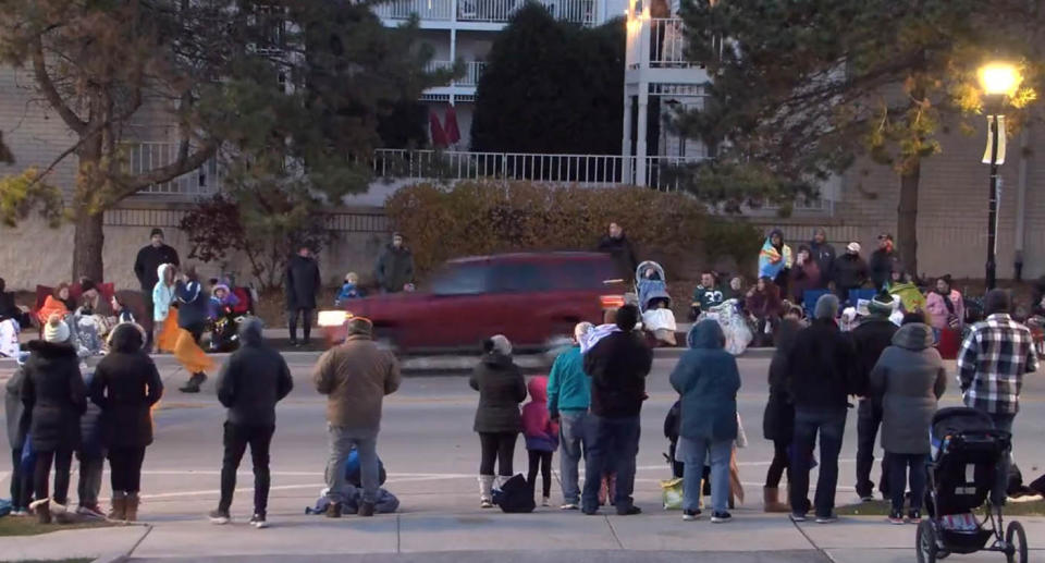 Screenshot from video showing red car speeding down street with people standing on the side of road.