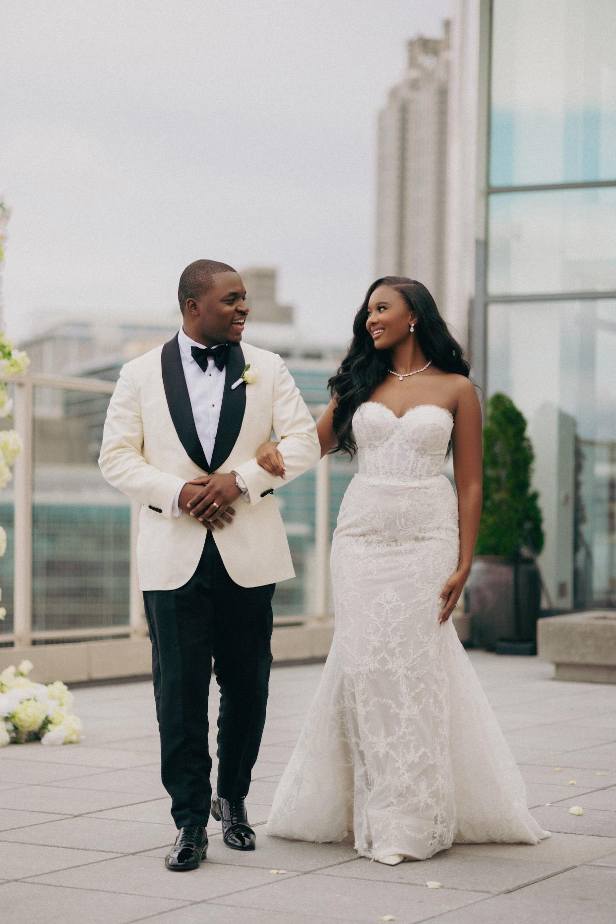 A bride and groom link arms and smile at each other on a rooftop in their wedding attire.