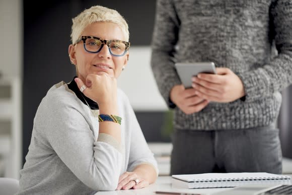 A mature woman sits at a desk in an office. A person stands in the background.