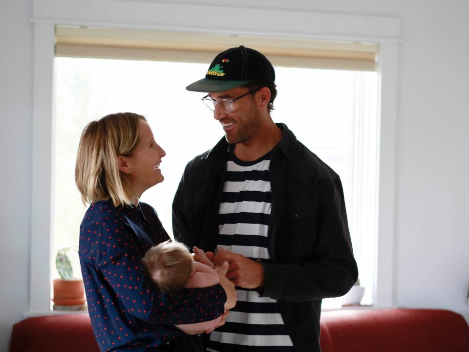 a man and a woman stand and smile at each other while holding their newborn.