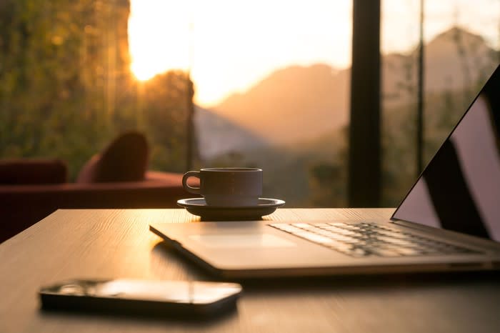 A laptop, smartphone, and cup of coffee sitting on a table overlooking a bank of windows.