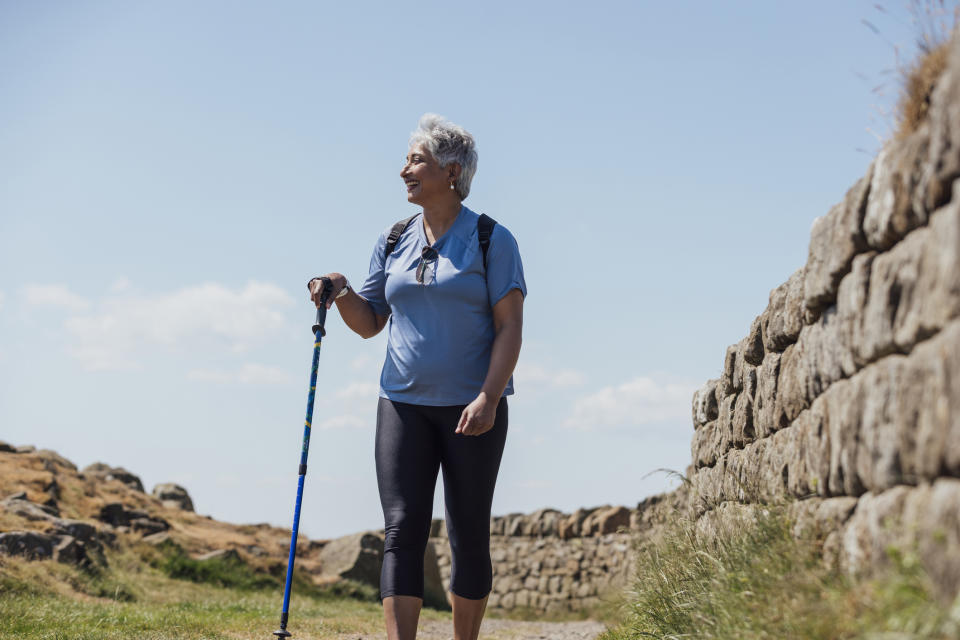 Older woman with short gray hair hiking along a stone wall, using a walking pole and carrying a backpack, smiling and dressed in active wear