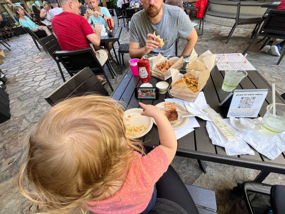 A toddler sitting on someone's lap pointing to a table full of food with someone eating across from her.