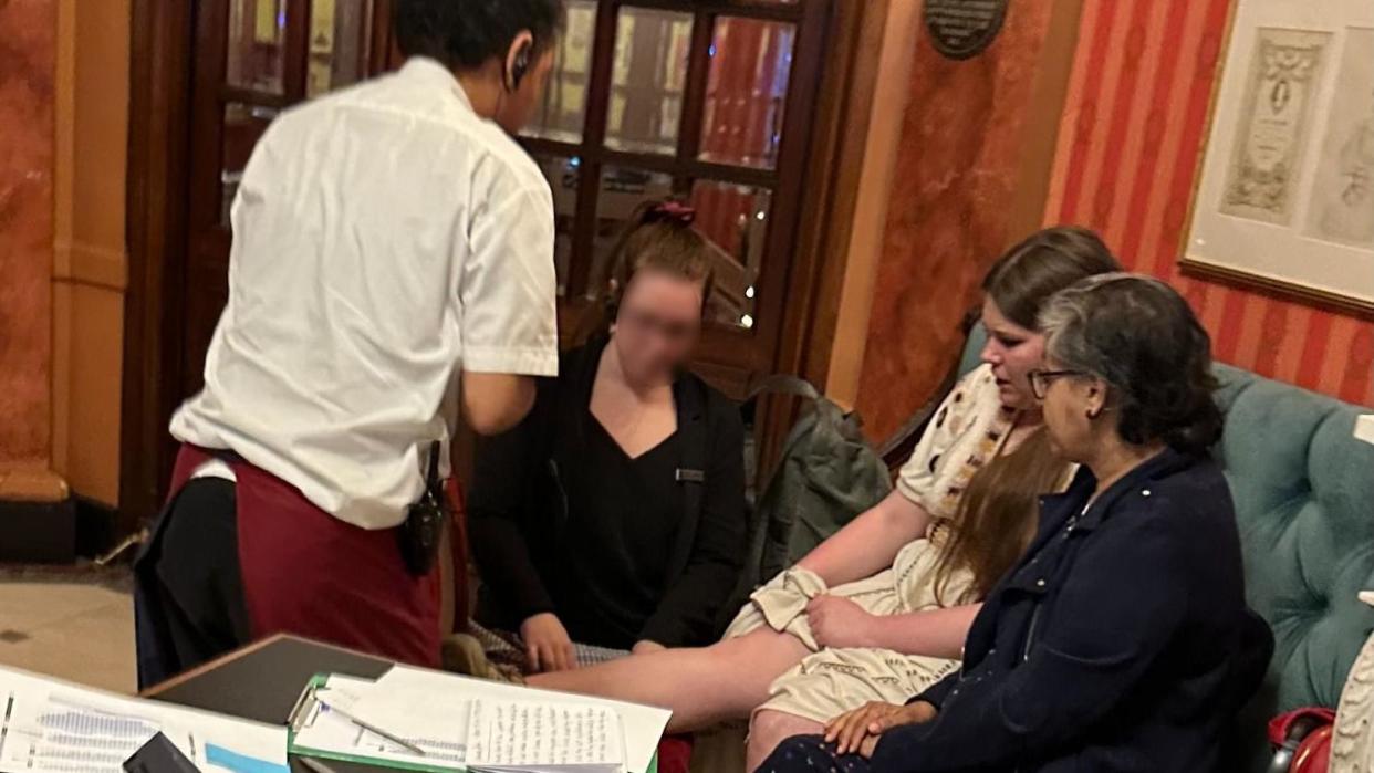 A woman sits with her leg elevated and an ice pack on it, surrounded by three people inside a theatre foyer.