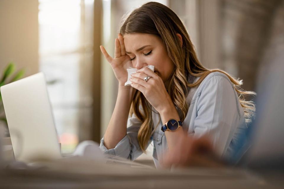 A woman holds a tissue to her nose while sitting in front of a laptop.