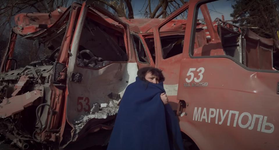 A woman hugs a blanket around herself, covering her mouth in front of a destroyed vehicle in "20 Days in Mariupol."