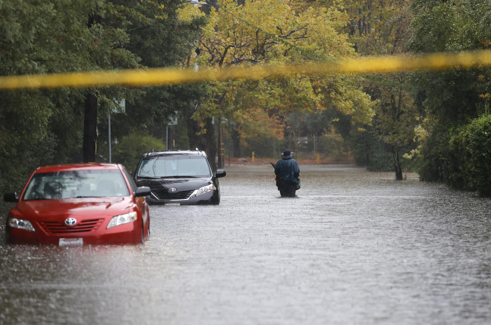 A pedestrian walks on a flooded street in the rain next to two partially submerged cars.