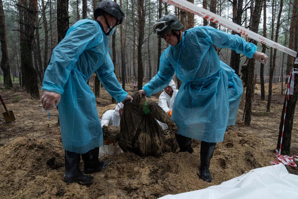 Two people in blue protective gear drag a body in a forest.