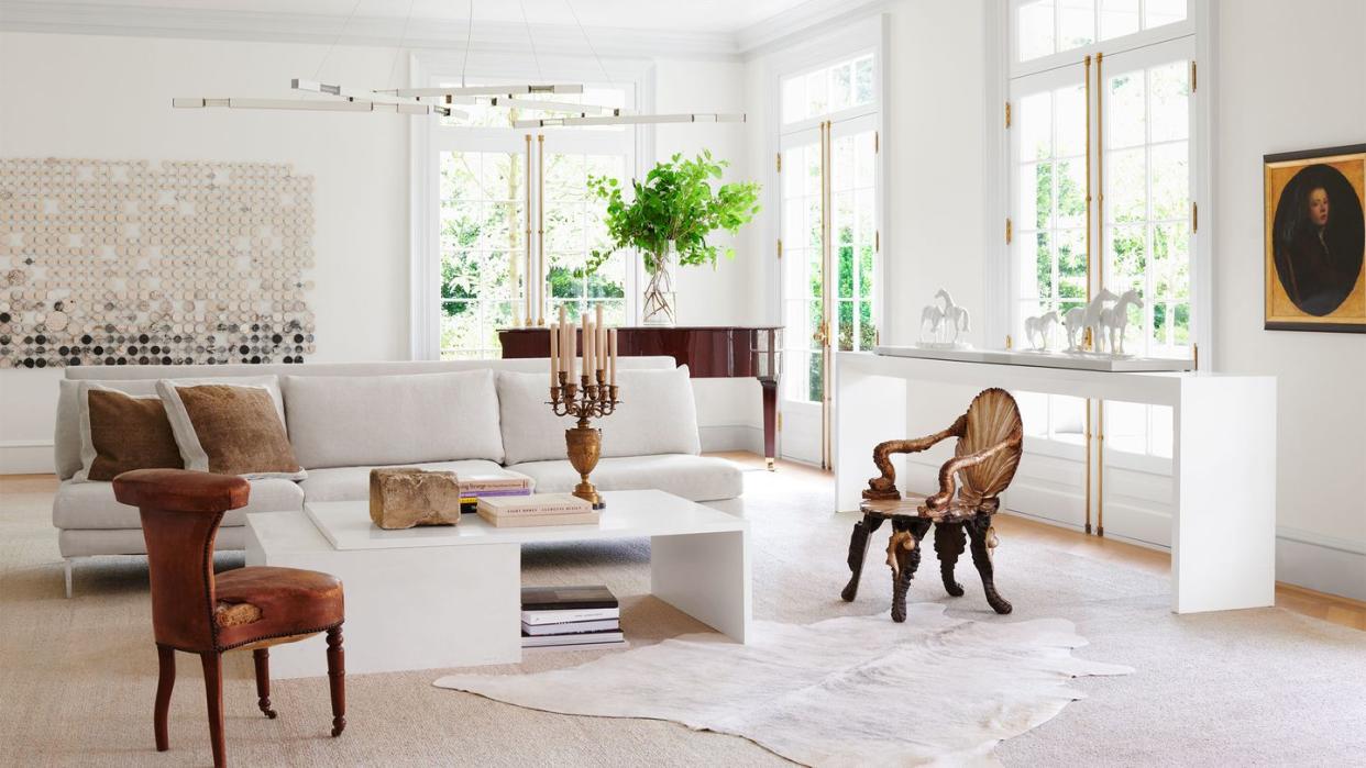 in a white living room is a white sofa, two side chairs, a white cocktail table with books and candelabra, white sideboard, a grand piano, white chandelier with brass arms, a large artwork on back wall, white carpet