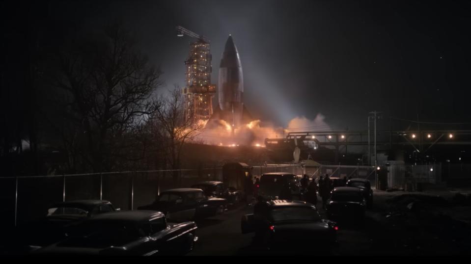 a rocket takes off at night time surrounded by dozens of 1950s-style cars