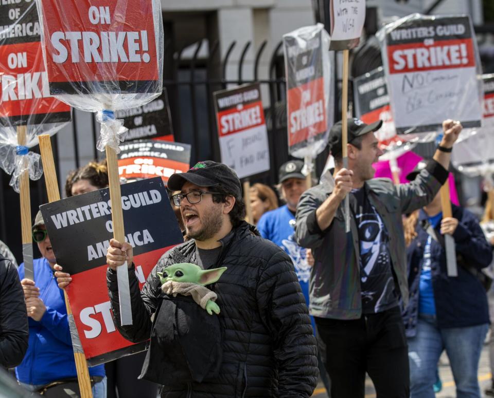 A man with a plush Baby Yoda carried against his chest holds a sign and hollers amid other striking writers