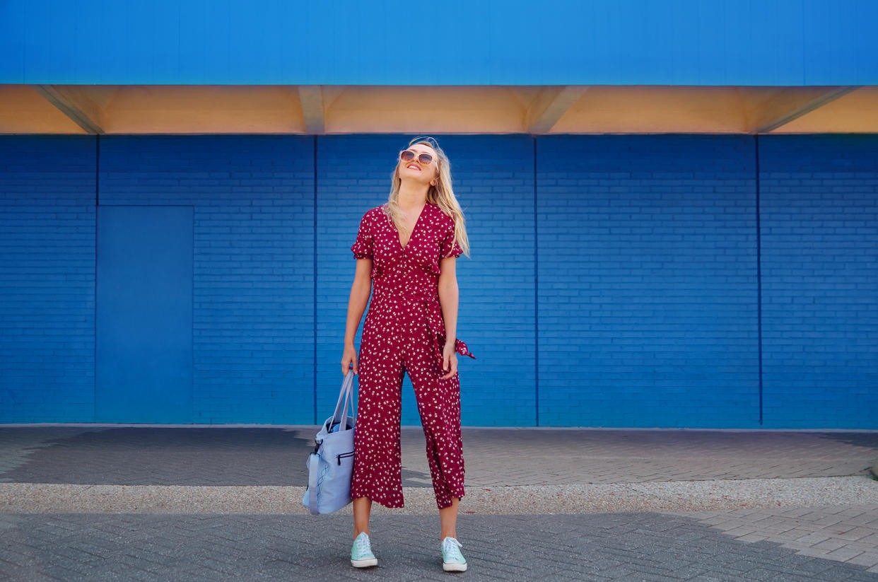 Pretty blond woman wearing burgundy jumpsuit and sunglasses while walking against the blue wall and holding cerulean tote bag