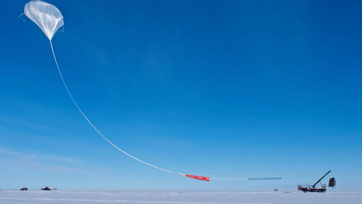  A large white scientific balloon launches into a blue sky from a flat, snow-covered patch of ground. 