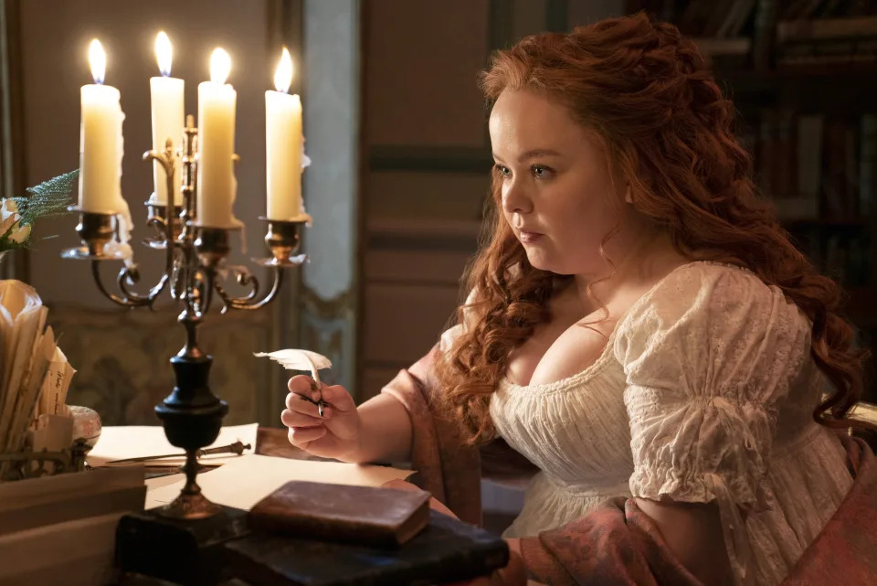 Nicola Coughlan is seated at a desk holding a quill pen next to a candle holder with lit candles in a scene from Bridgerton