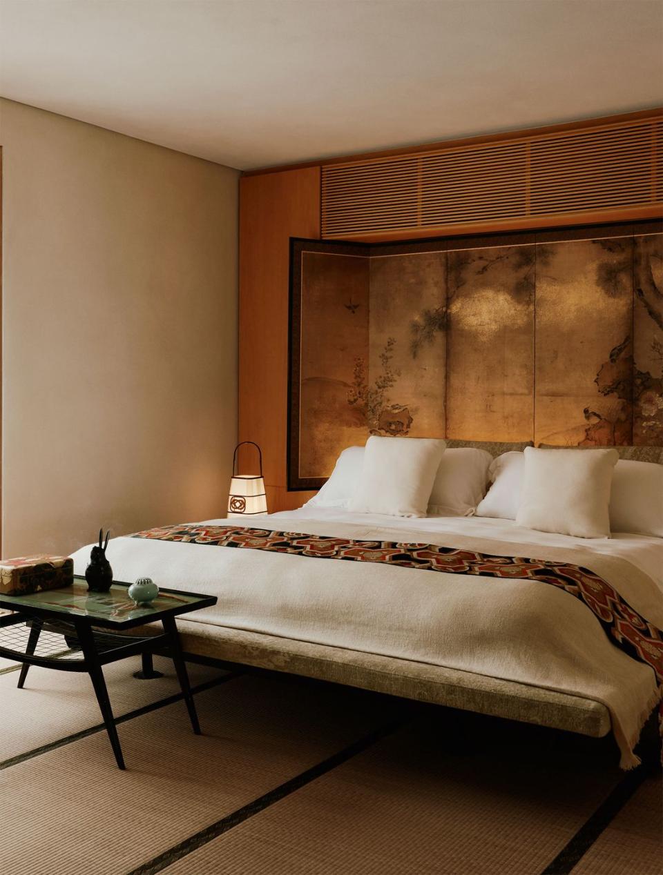 a platform bed with a fabric frame, white bedding with a printed sash at the foot, tatami floor mat, small table at foot of bed, built in wood cabinets behind bed with a japanese screen, paper lamp