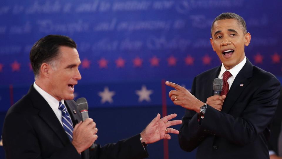 mitt romney and barack obama, both wearing black suits and ties, speak simultaneously into microphones on a stage during a debate
