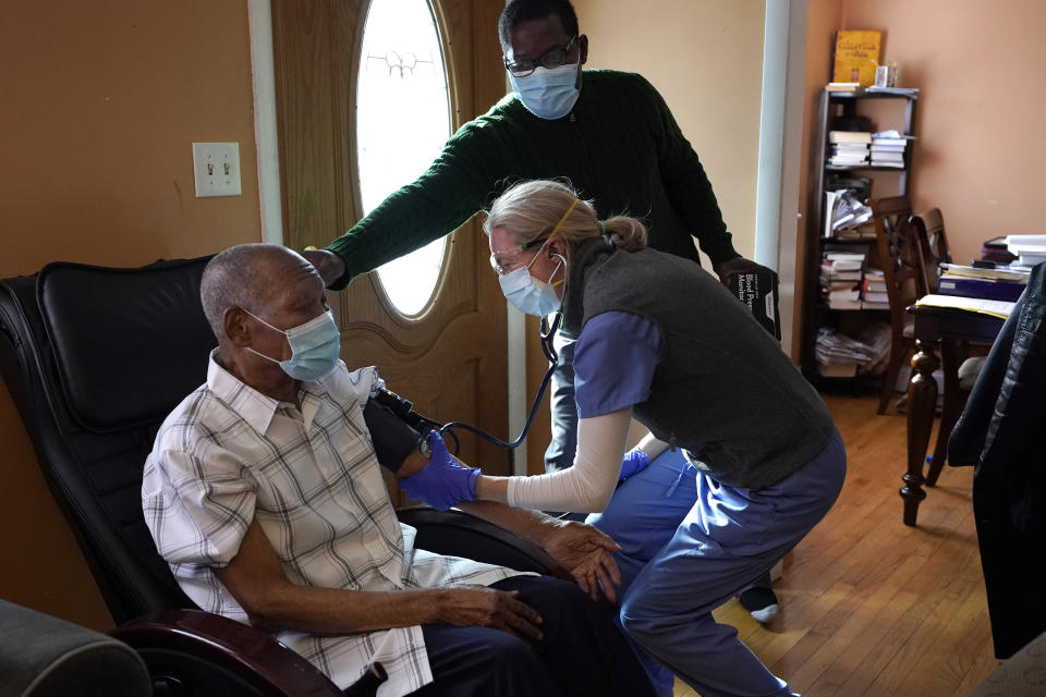 Edouard Joseph, 91, left, has his blood pressure taken by geriatrician Megan Young, right, as Joseph's son, Edouard F. Joseph, top, offers support after his father received a COVID-19 vaccination, Thursday, Feb. 11, 2021, at his home in the Mattapan neighborhood of Boston. Millions of U.S. residents will need COVID-19 vaccines brought to them because they rarely or never leave home. Doctors and nurses who specialize in home care are leading this push and starting to get help from state and local governments around the country. (AP Photo/Steven Senne)