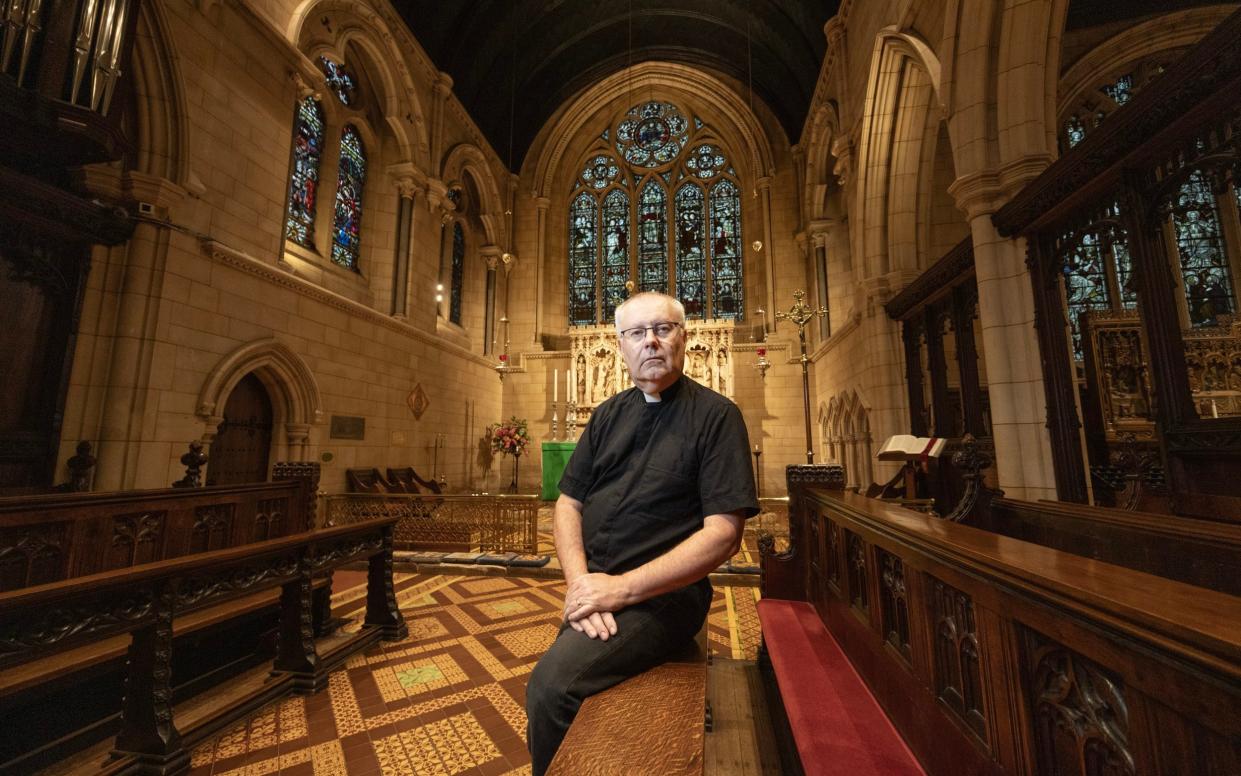 Father Brandes perches on a pew in the church, with its high stained-glass altar window behind him