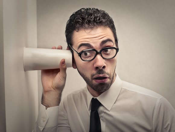 A man in glasses holds a cup to his ear and a wall attempting to listen to what's  on the other side.