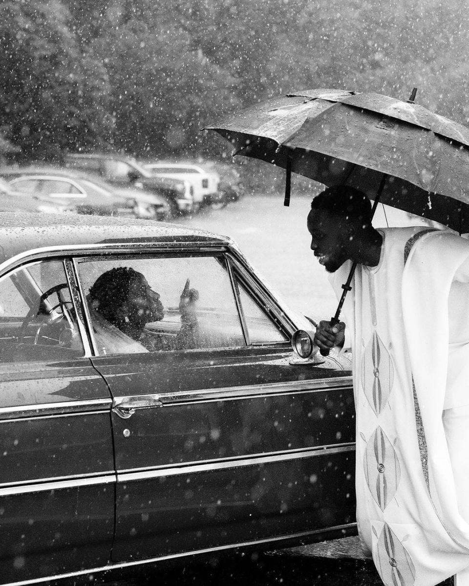 A groom holding an umbrella leans down to a car window to greet this bride.