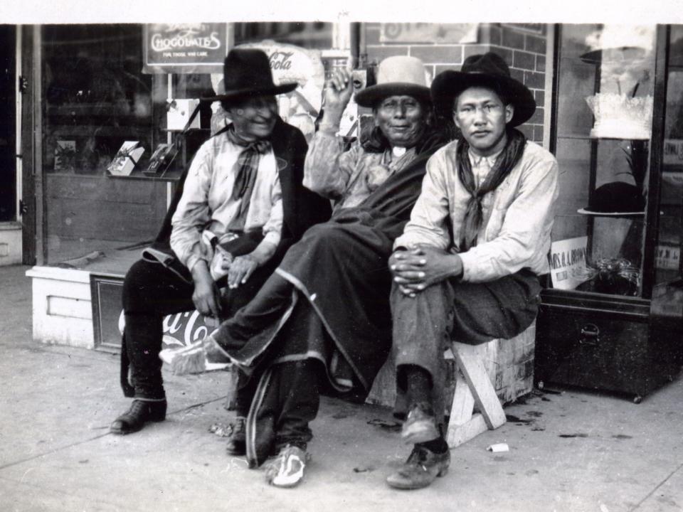 Postcard features a photograph of three of unidentified men of the Osage Nation as they sit on a bench in front of shop, Pawhuska, Oklahoma Territory, circa 1918 - 1919.