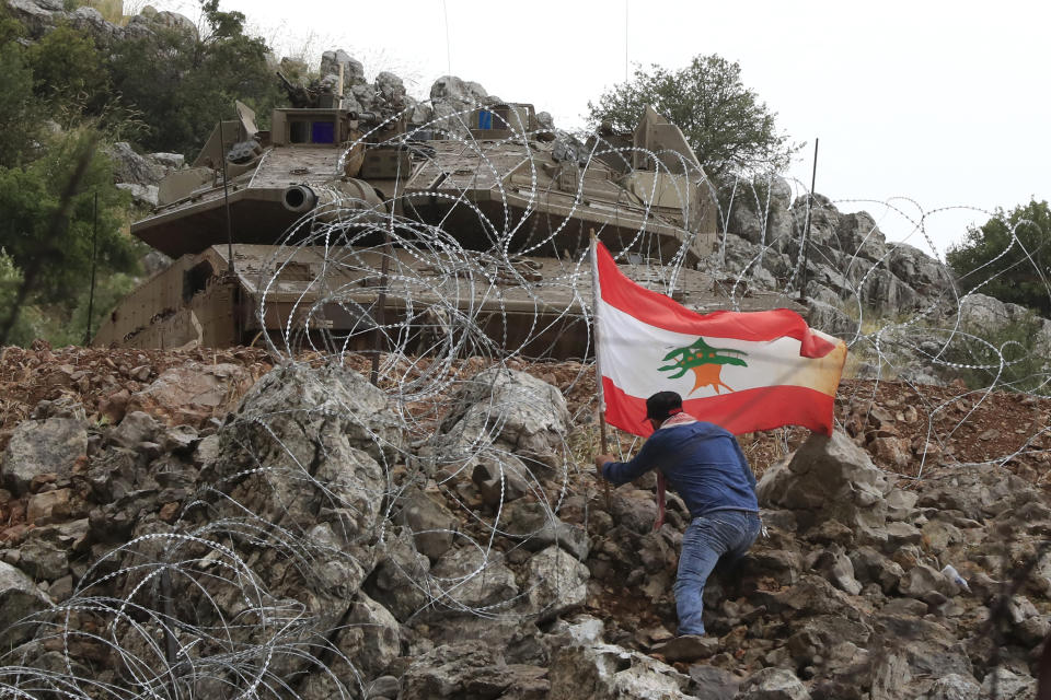 A Lebanese protester sets a Lebanese flag under a Israeli Merkava tank in the disputed Kfar Chouba hills along the border, in south Lebanon, Friday, June 9, 2023. Israeli soldiers fired tear gas to disperse scores of protesters who pelted the troops with stones along the border with Lebanon Friday, leaving some Lebanese demonstrators and troops suffering breathing problems. (AP Photo/Mohammad Zaatari)