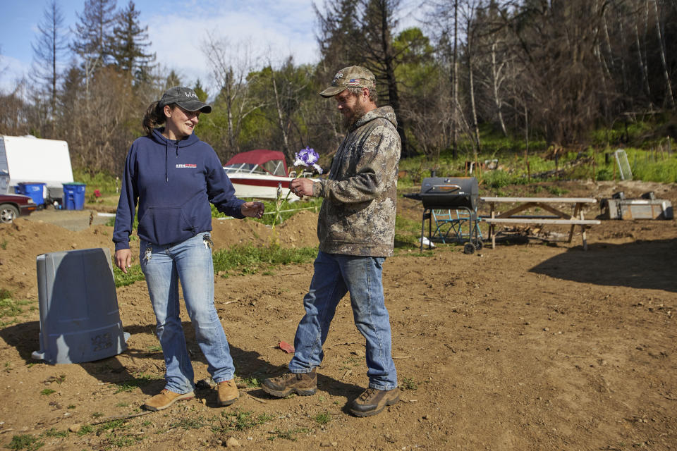 Tye Small hands his wife Melynda a flower he found while clearing debris from their property in Otis, Ore., on Thursday, May. 13, 2020. They are standing where their house was before the Echo Mountain fire destroyed it. The Smalls have dedicated their time to helping the small town on the Oregon coast recover from the devastating fire that destroyed 293 homes. Experts say the 2020 wildfire season in Oregon was a taste of what lies ahead as climate change makes blazes more likely and more destructive even in wetter, cooler climates like the Pacific Northwest. (AP Photo/Craig Mitchelldyer)