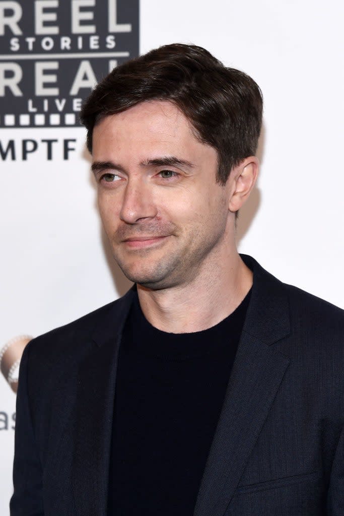 Man in a dark blazer at an event, looking to the side, with event logos in the background