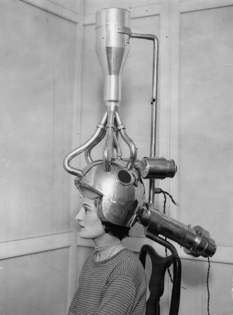 A seated woman underneath a metallic head covering that looks like armor, with metallic cables emerging from it and extending toward the ceiling or behind her