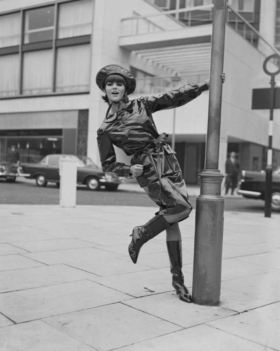 Model Jackie Bowyer swings on a lamppost wearing a black oilskin wet-weather outfit from Mary Quant, of matching coat and hat with shiny, high-heeled boots, London, 15th October 1963 (Getty Images)