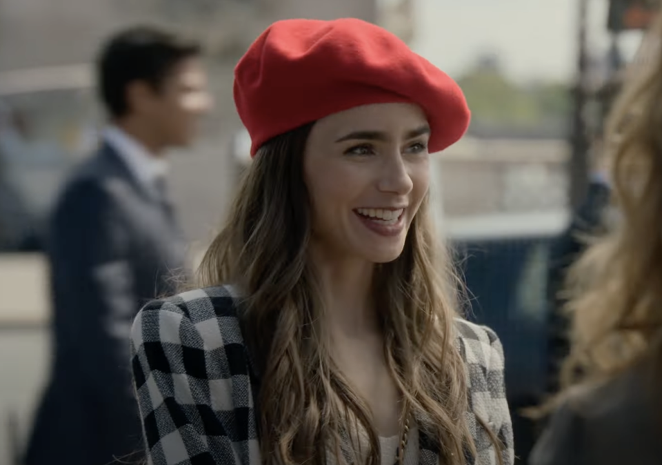 Lily Collins smiles while wearing a red beret and a checkered coat, interacting with another person outdoors in a city setting