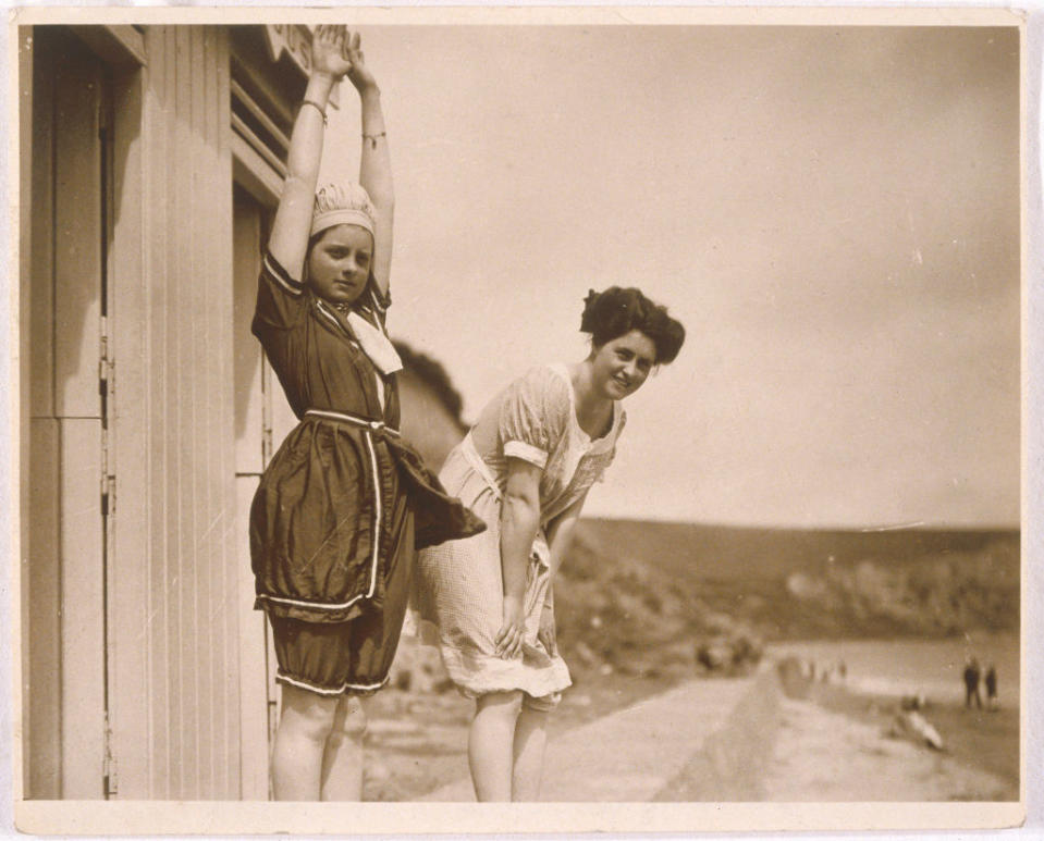 Two vintage-era women posing beside a building, one with arms raised, the other with hands on hips