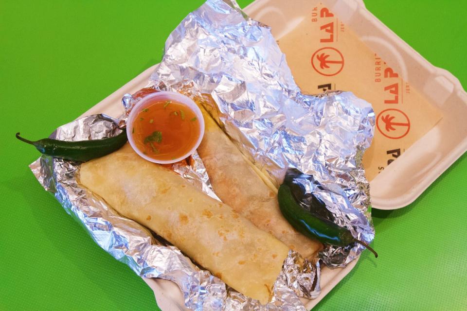 Two burritos in a takeout container with two peppers and red salsa.