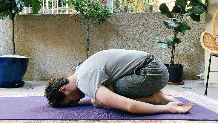 Man kneeling on a yoga mat while leaning forward with his arms alongside his body in Child's Pose
