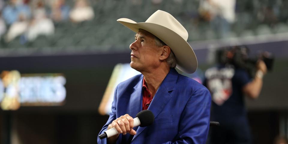 Wearing a tan cowboy hat, Texas Governor Greg Abbott lowers a microphone toward his chest while listening to the national anthem at a World Series baseball game held in Arlington, Texas.