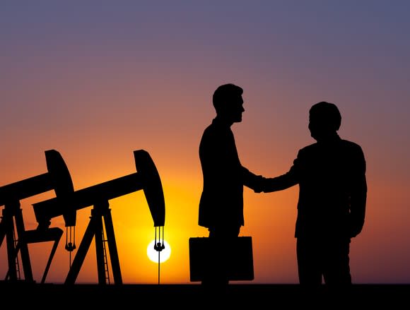 The silhouette of two people shaking hands with oil pumps in the background.
