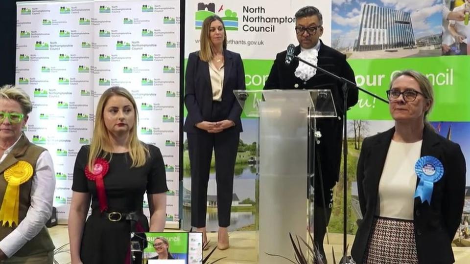 High Sheriff dressed in black and white uniform speaking into a microphone with candidates wearing rosettes