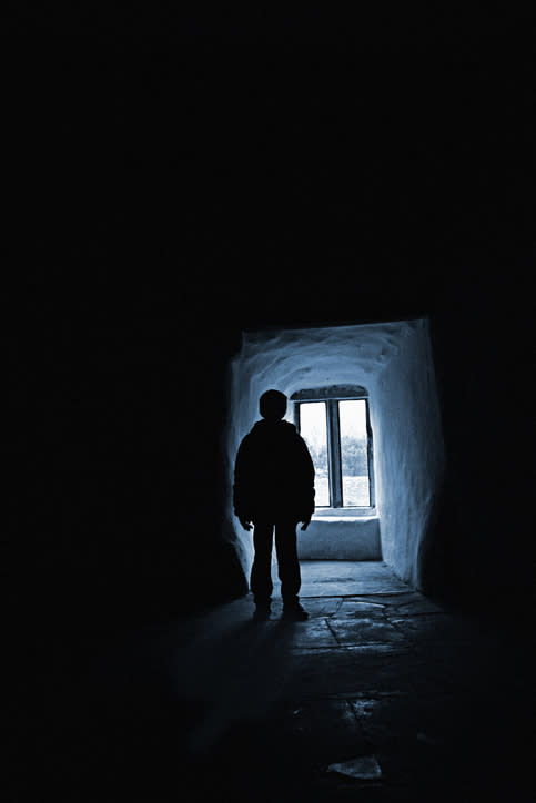 Silhouette of a person standing in a narrow, dimly lit hallway, facing a small window with natural light streaming in