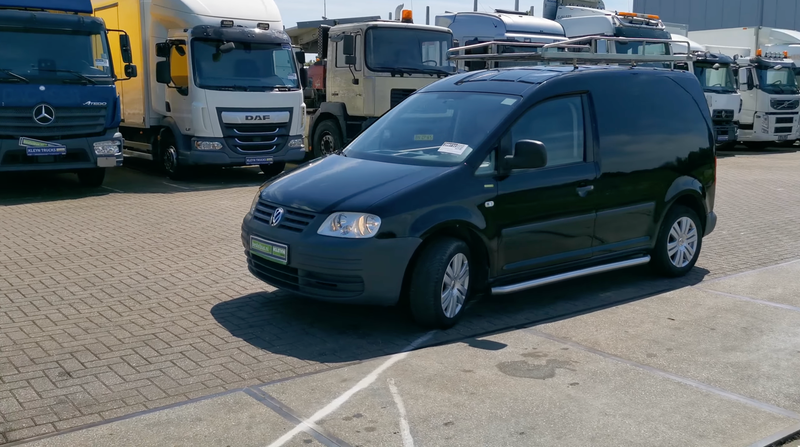 A black VW Caddy Maxi panel van in black in front of a bunch of euro semi trucks.