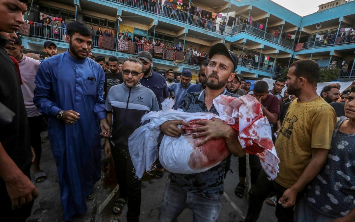 The child's body is wrapped in a blood-stained sheet and the father holds it in the middle of a crowd of onlookers