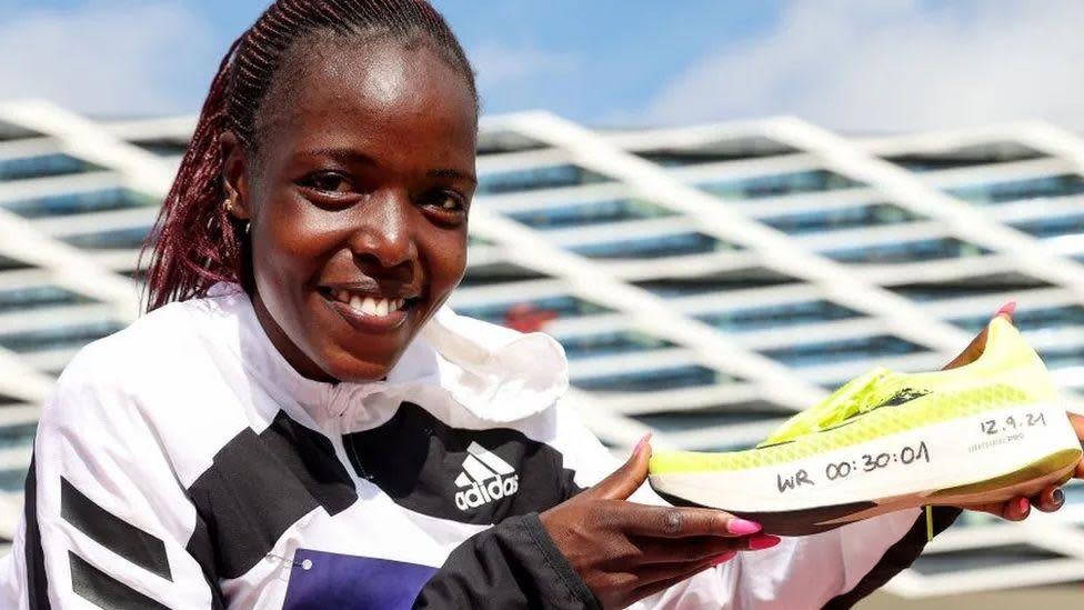 Agnes Jebet Tirop holding a running shoe with the following written on the side by hand: WR 00:30:01