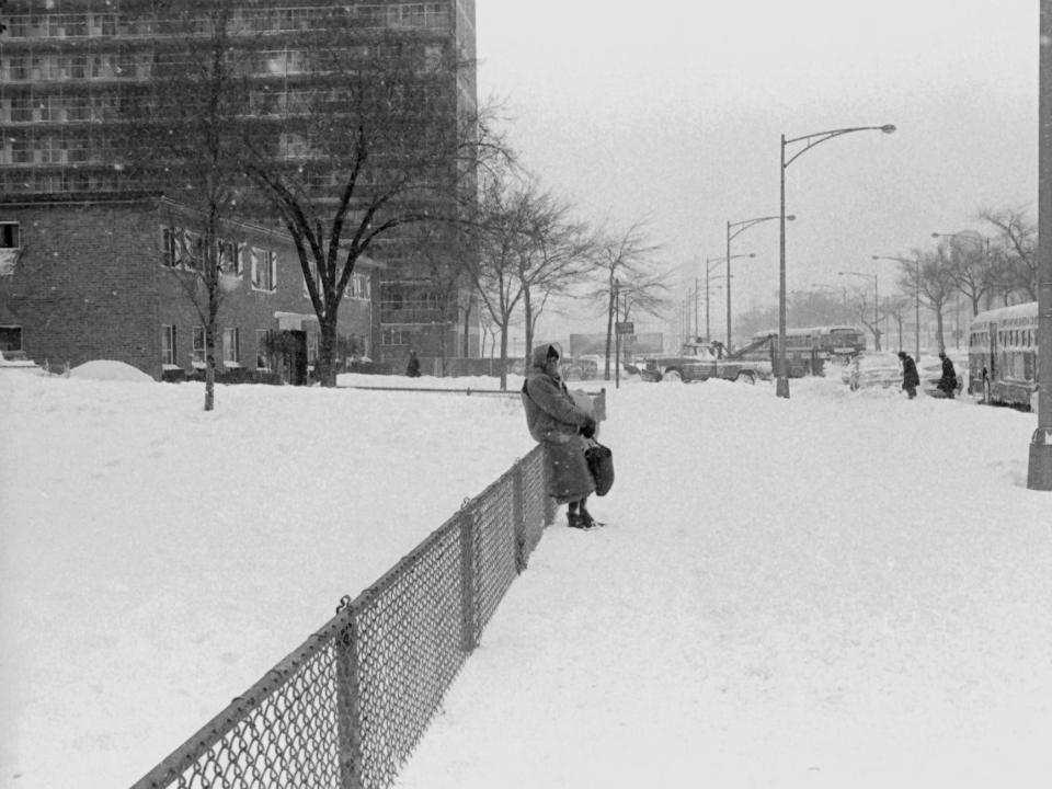 On a snow covered sidewalk, a woman waits for a Chicago Transit Authority (CTA) bus during a winter blizzard in Chicago, IL, January 1967. Snow is piled high all along the streets.