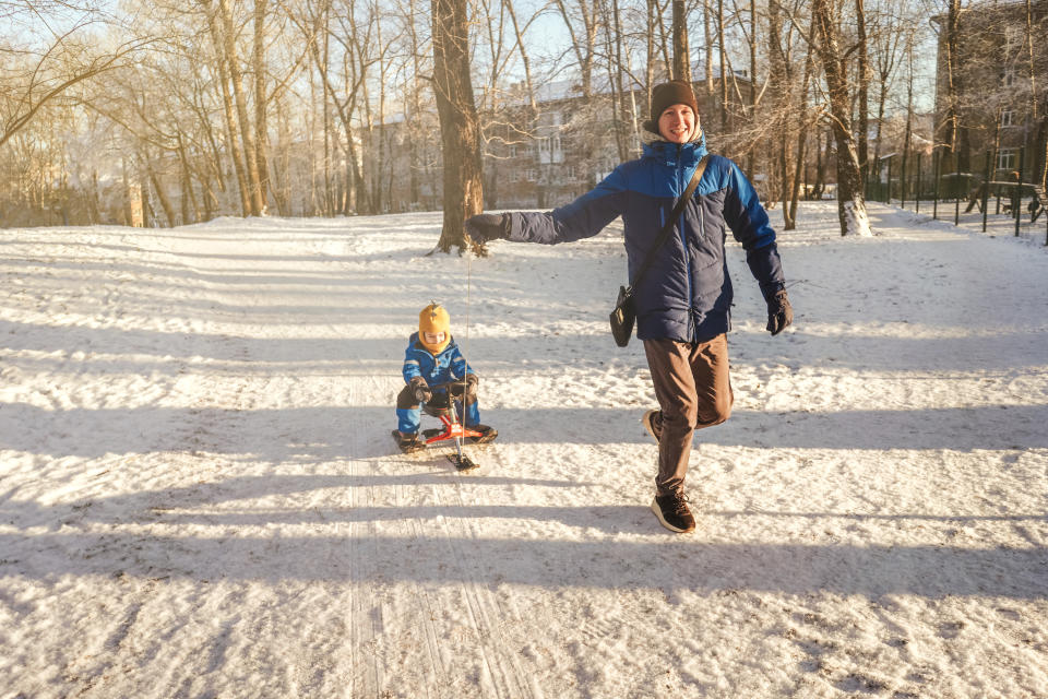 A man run and rolls a small child boy on a snow racer through the snow from the mountain in the park.