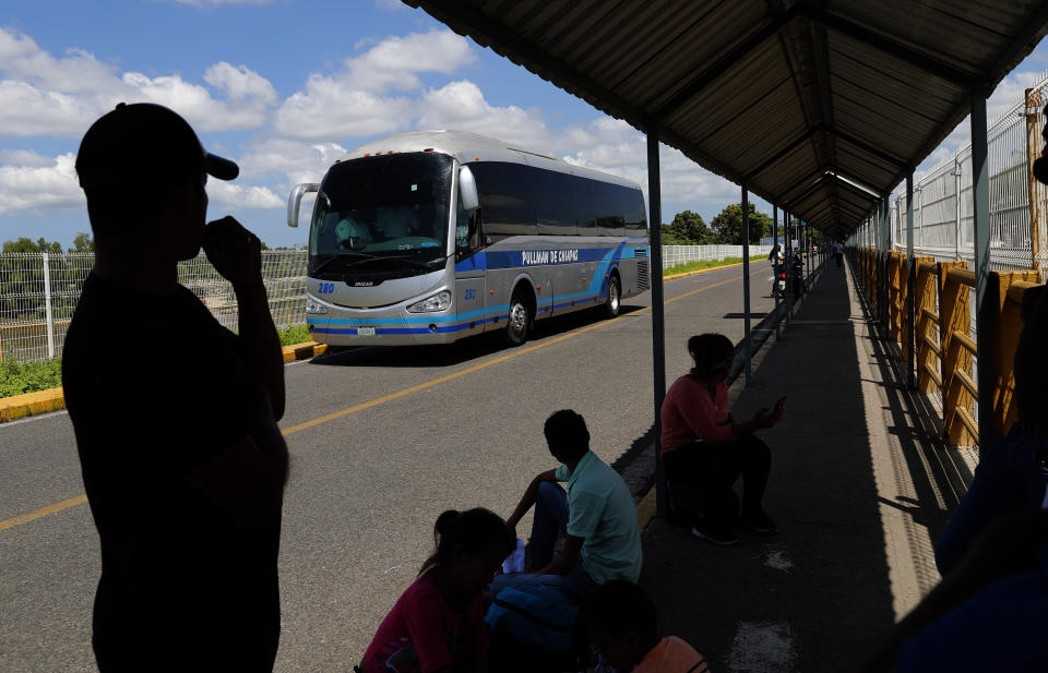 Honduran migrant Jose Maria Dubon Gutierrez, left, waits at a border crossing between Guatemala and Mexico for his family's turn to show Mexican immigration officers their documents after securing legal permission to stay in Mexico's southern states, near Ciudad Hidalgo, Mexico, Thursday, June 6, 2019. Behind, a bus passes, which local authorities say carry Central American migrants back to Guatemala, one day after Mexican police and immigration agents blocked the advance of about 1,000 Central American migrants who were walking along a highway. (AP Photo/Marco Ugarte)