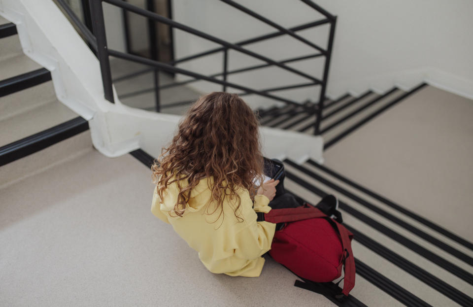 A person with curly hair, wearing a yellow hoodie, sits on stairs, looking at a tablet or phone. A red backpack is next to them. Scene appears candid and casual