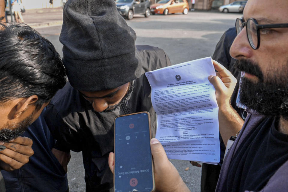 Caritas volunteer Giulio Escalona, right, holds a phone from which a Bengalese-speaking cultural mediator explains to a group of Bangladeshi migrants the terms of their expulsion order in front of Salerno's railway station, southern Italy, Monday, Oct. 23, 2023, where they were brought after being rescued from the sea on Oct. 6 by the humanitarian ship Geo Barents in the Mediterranean Sea some 30 miles off Libya. Badly advised by relatives and friends, misled by insufficient official information or poor translation services, many can end up in legal limbo for years, cut off from any government aid. (AP Photo/Paolo Santalucia)