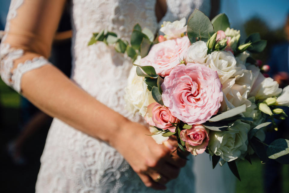 A close-up of a bride holding a bouquet of pink and white roses. The bride's lace dress sleeve is partially visible.